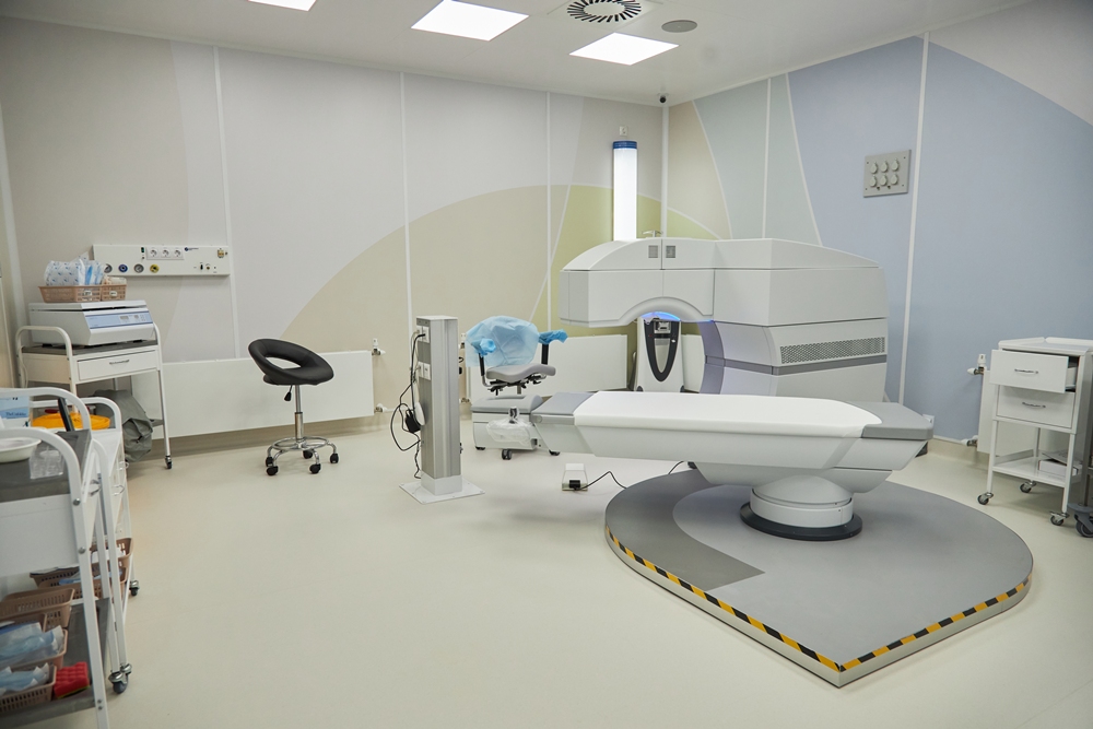Medical imaging room featuring a large CT scanner and a sliding patient table with surrounding medical equipment on carts and chairs.