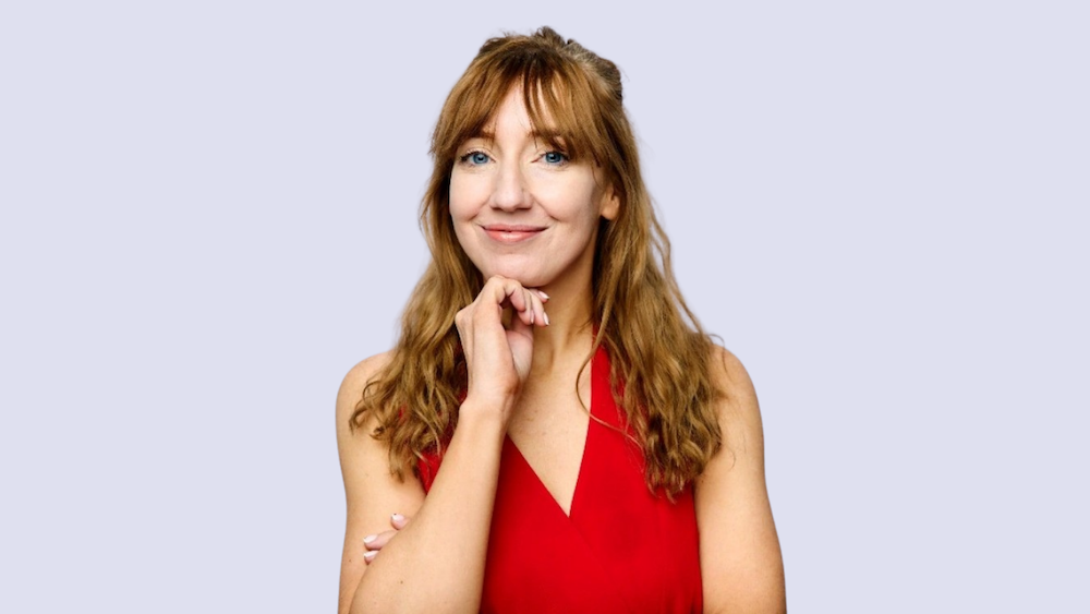 Woman with wavy red hair in a red sleeveless top, smiling softly and resting her chin on her hand against a pale lavender background.