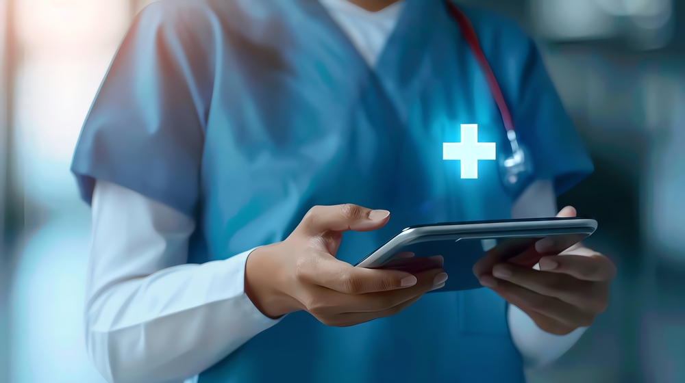 Healthcare professional in blue scrubs with a stethoscope, using a tablet in a clinical setting.