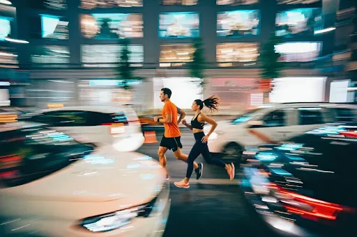 Two runners, a man in an orange shirt and a woman in black, sprint through a busy city street as cars blur past.